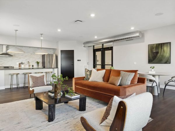 Modern open-plan living room and kitchen with a brown sofa, bar stools, and dining table in neutral tones.