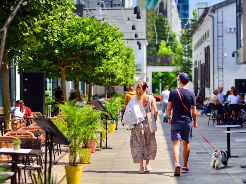 A couple walks a dog down a sunny outdoor café-lined street with people dining and green trees near The Espen Reisdences