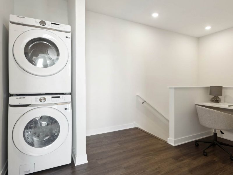An in-home laundry area at The Espen Residences in Oakdale, MN, featuring a stacked washer and dryer next to a small workspace with wood-style flooring and recessed lighting.