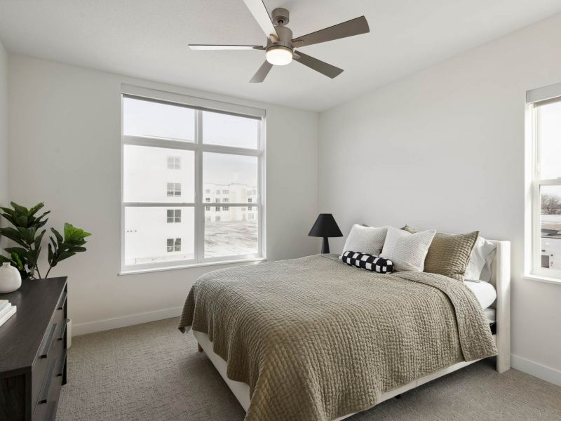 A bright bedroom at The Espen Residences in Oakdale, MN, featuring a large window, neutral bedding, a ceiling fan, and clean modern finishes.