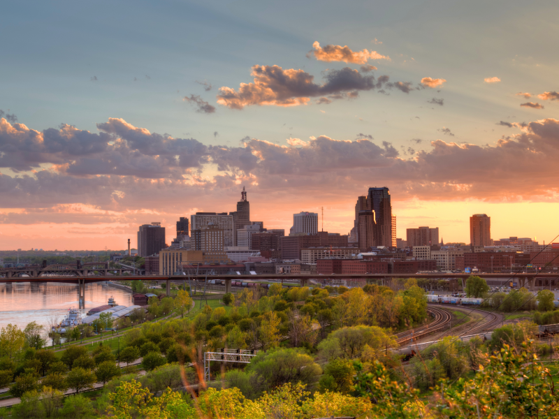 Scenic view of the downtown St. Paul, MN skyline at sunset, featuring city buildings, bridges over the Mississippi River, lush greenery, and colorful clouds illuminated by the evening sky.