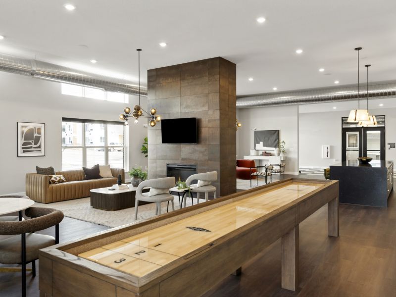 Modern community room at The Espen Residences in Oakdale, MN, featuring a shuffleboard table, comfortable lounge seating, a fireplace, contemporary lighting, and a kitchen area.