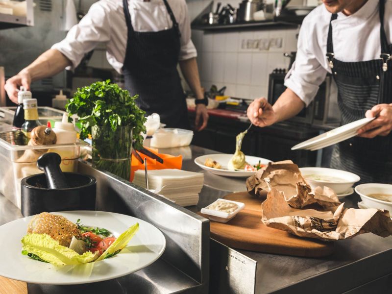 Two chefs in a kitchen preparing and plating dishes with fresh ingredients and greens on the counter near The Espen Residences